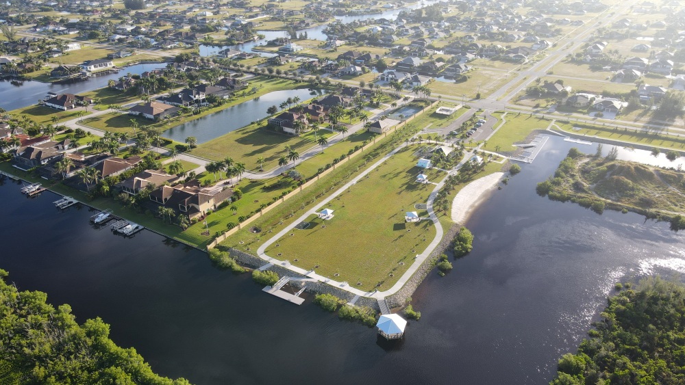 Aerial view of Tropicana Park on the water, showing a walking path, a sandy beach, picnic shelters, boat docks and a covered observation deck at the end of a pier, with homes and canals around the park.